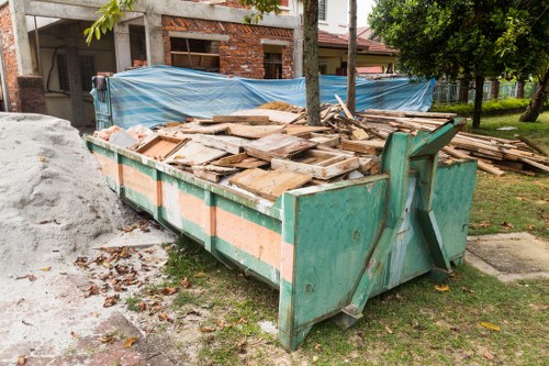 A large, rectangular metal skip painted in faded green, situated on a grassy area next to a residential property, filled with an assortment of wooden debris, including planks, panels, and broken pieces of timber. The timber appears weathered, with some pieces showing signs of age such as rough textures, splinters, and uneven cuts, and is piled haphazardly inside the skip. Surrounding the skip, there are scattered dead leaves and small twigs on the grass, with a pile of discarded building materials, possibly concrete or plaster, nearby on the left. In the background, a partially constructed or renovated brick building with scaffolding and blue tarpaulin is visible, along with a row of trees and a paved footpath. The scene suggests a waste clearance operation in a residential area near Aldershot, with House Clearance Aldershot providing rubbish removal services for construction or renovation debris.