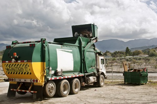 A large green and yellow skip loader truck parked on a gravel surface outdoors, with a closed green waste container to its right, filled with mixed waste materials, including wooden planks and other debris. The truck features a prominent yellow caution sign at the rear and is positioned in front of a landscape with mountains and a cloudy sky, suggesting an outdoor rubbish collection or clearance site typical of waste removal services near Aldershot. The scene captures the vehicle's side view, highlighting its substantial size, robust wheels, and operational design for rubbish collection, aligning with services offered by House Clearance Aldershot in the Hampshire area.
