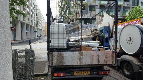 A flatbed delivery truck parked on a city street with a metal mesh side panel, carrying a load of assorted rubbish and discarded items. The visible debris includes white and blue furniture components, wooden panels, and various unidentifiable waste materials. The truck’s surface shows signs of rust and dirt, with a yellow and black license plate reading 'NX12 GHA.' In the background, modern multi-storey residential buildings with white exteriors and balconies line the street, and a pedestrian can be seen walking across a crosswalk. The scene suggests a rubbish collection or house clearance operation typical of services offered by House Clearance Aldershot, situated in the Aldershot area. The environment is urban, with a paved sidewalk and passing traffic, indicating a typical residential or commercial area in the vicinity of Aldershot.