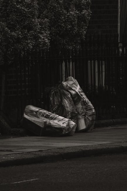 Two large black garbage bags and a cardboard box are placed on the edge of a sidewalk next to a black metal fence. The bags appear to be filled with waste, with their glossy, crinkled surfaces reflect
